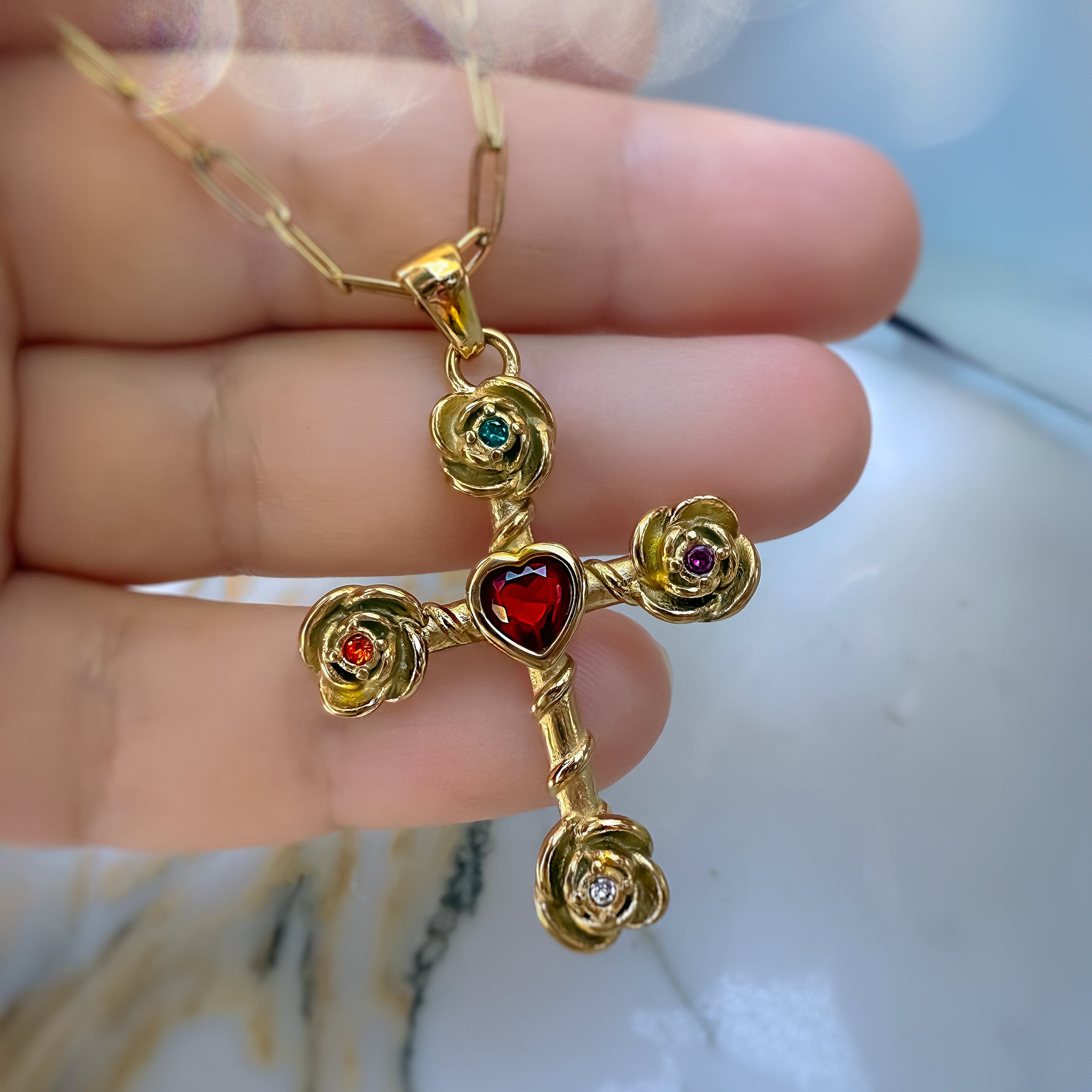 Gold cross pendant with red heart and gemstones held by a hand on a marble surface