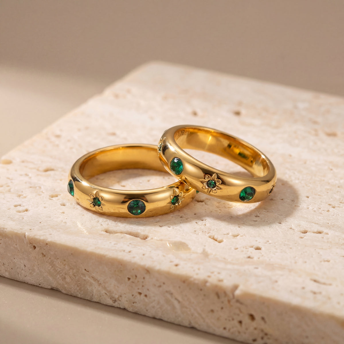 Two gold rings with emerald stones on a beige stone surface