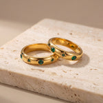 Two gold rings with emerald stones on a beige stone surface