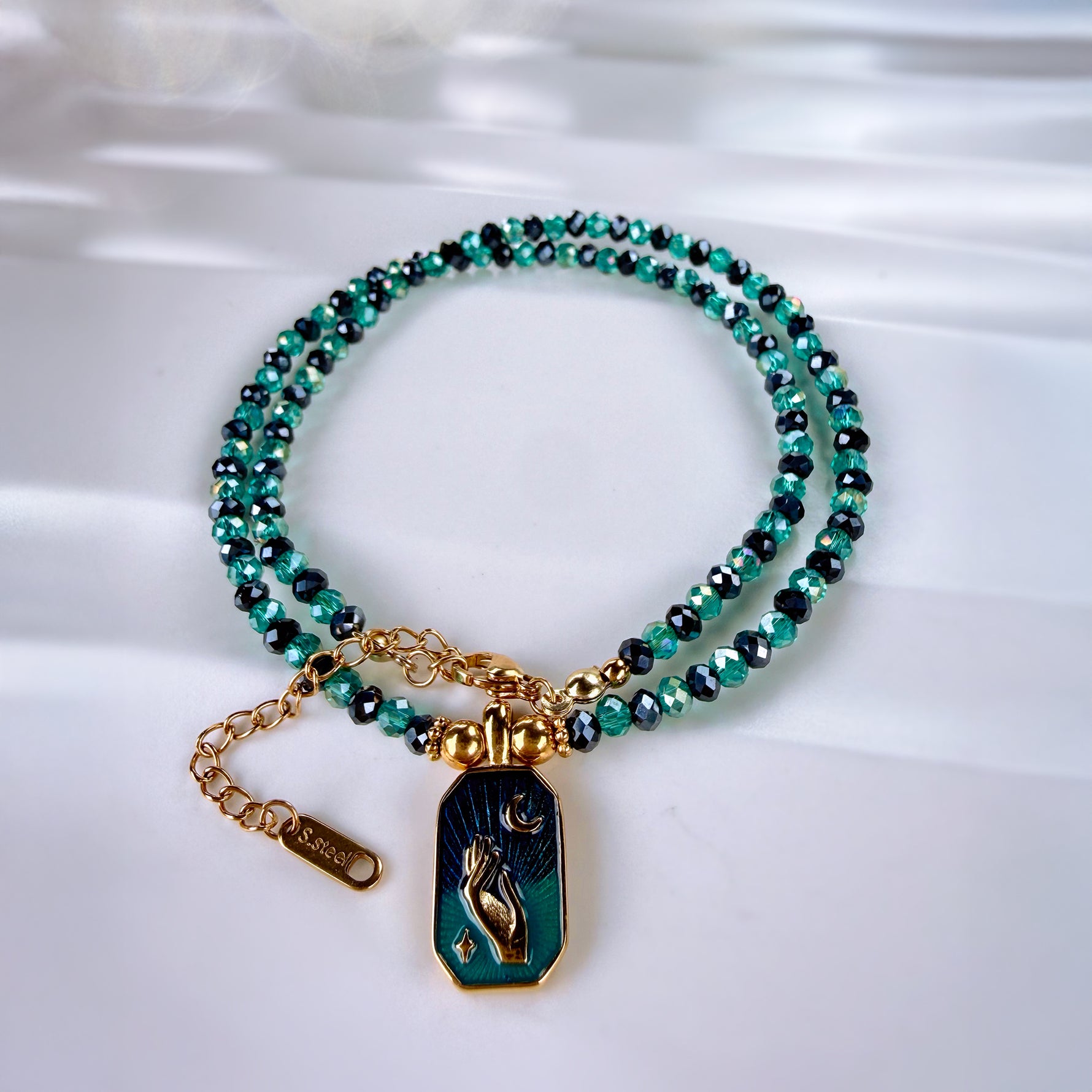 A double-strand beaded bracelet with teal and black faceted beads, gold-tone chain links, and a coffin-shaped pendant featuring a gold skeleton figure on a dark teal background. The brand name 'SARABE' is visible on the gold clasp tag.