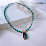 A beaded necklace with small turquoise and dark blue-green beads, featuring a gold-tone pendant with a blue and green mermaid design, displayed on a white marble block.