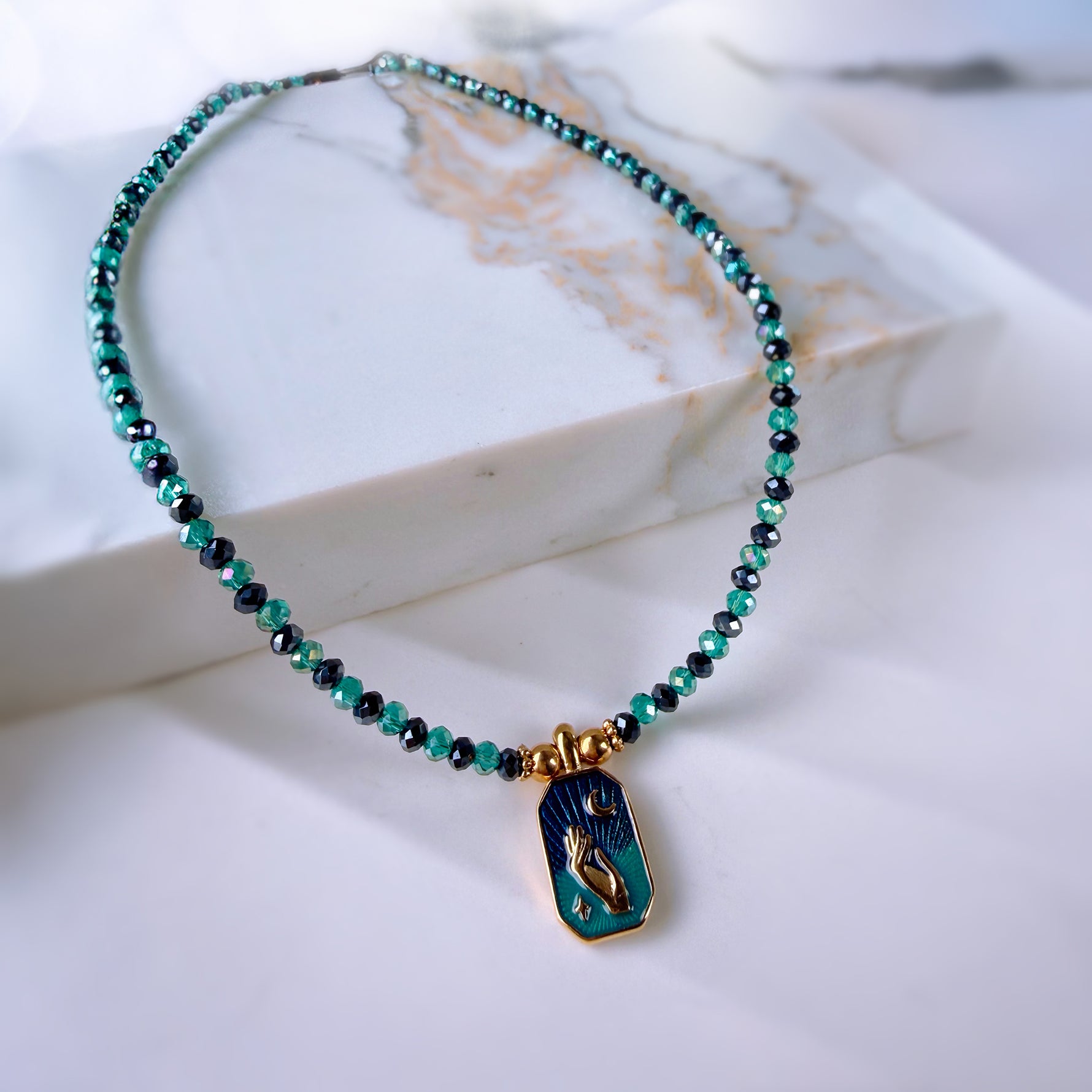 A beaded necklace with small turquoise and dark blue-green beads, featuring a gold-tone pendant with a blue and green mermaid design, displayed on a white marble block.
