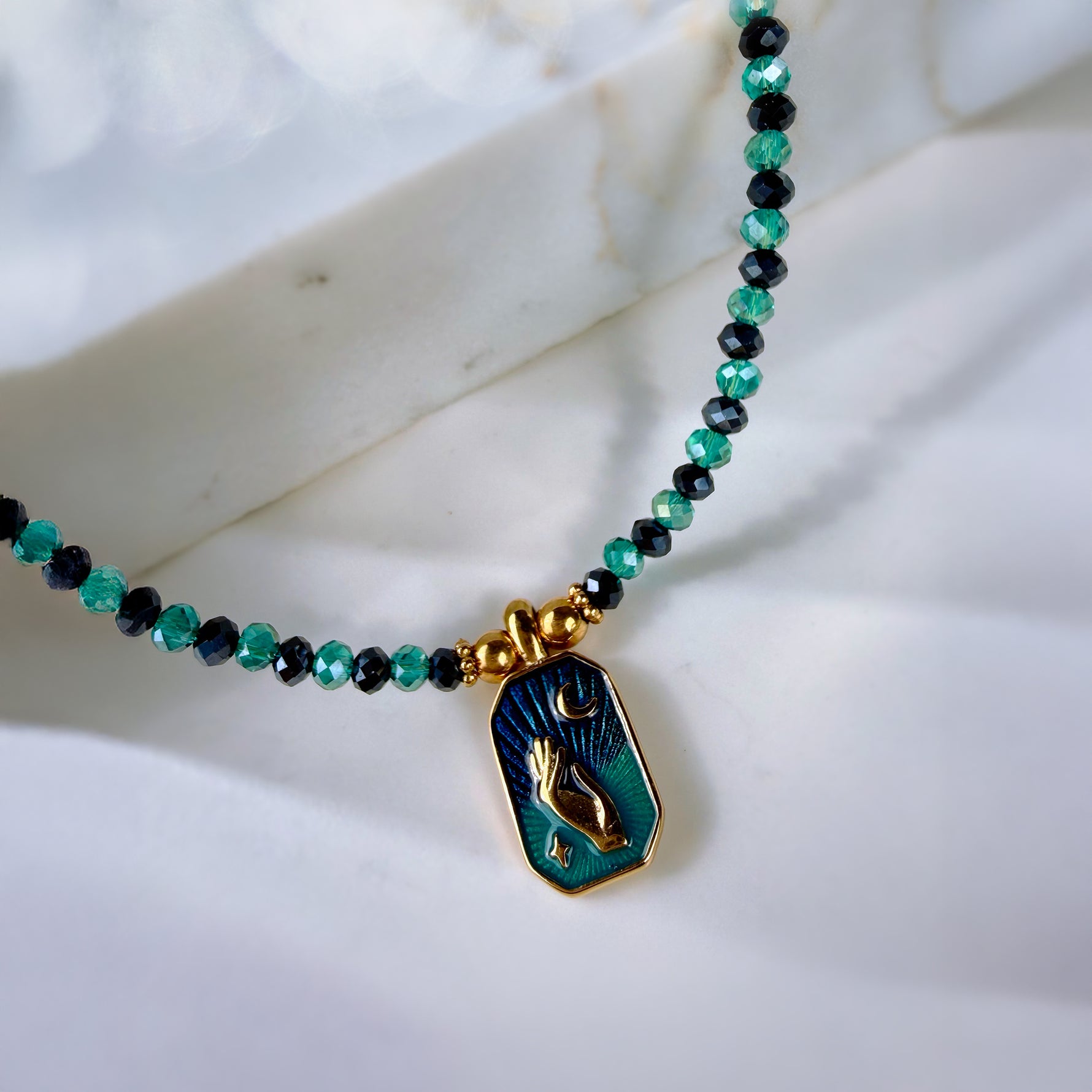 A beaded necklace with green and black faceted beads and a gold-toned pendant featuring a teal and green religious icon, displayed on a white marble surface.