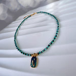 A beaded necklace with small green and black faceted beads, gold-toned clasp, and a dark blue-green pendant featuring a gold-toned figure, displayed on a white textured surface.