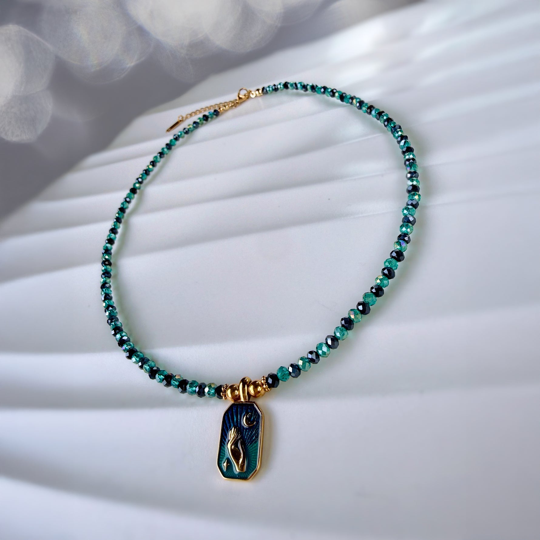 A beaded necklace with small green and black faceted beads, gold-toned clasp, and a dark blue-green pendant featuring a gold-toned figure, displayed on a white textured surface.