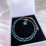 A beaded necklace with iridescent green and blue faceted beads, gold-tone chain links, and a small rectangular enamel charm, displayed inside an open white jewelry box with a black velvet insert.
