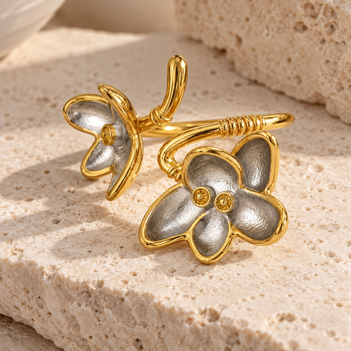 A pair of gold and silver flower-shaped cuff rings displayed on a textured beige stone surface.