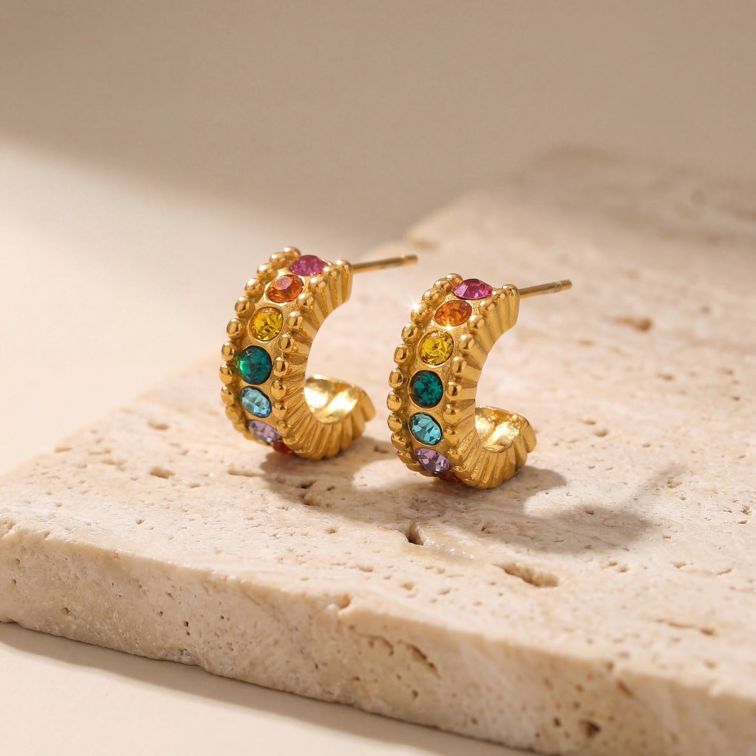 Gold hoop earrings with colorful gemstones on a beige stone surface