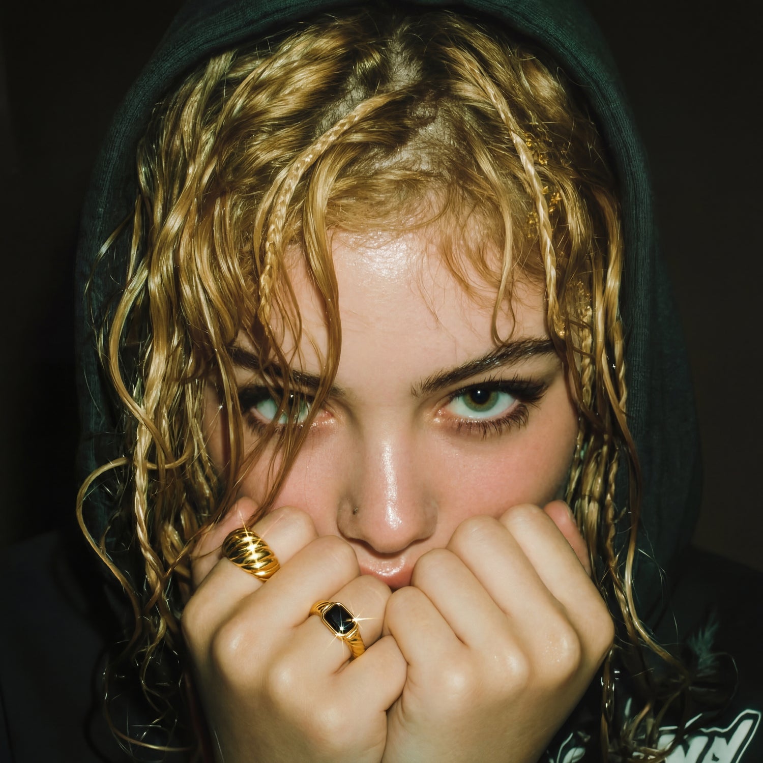 Person with wet hair wearing a dark hoodie, holding hands near face.