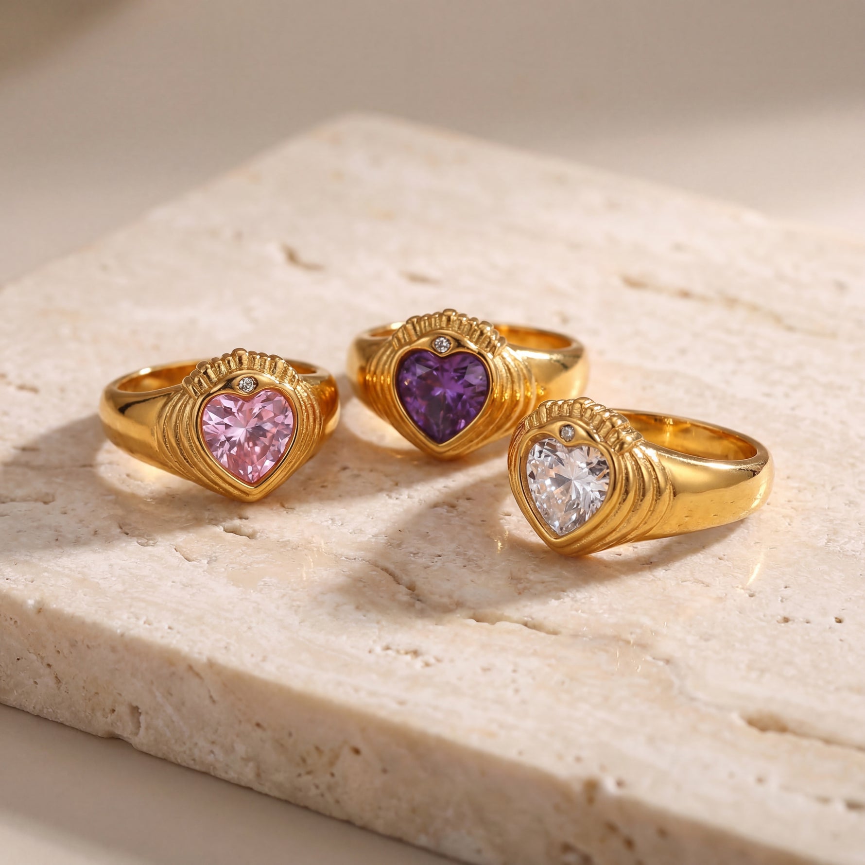 Three gold heart-shaped rings with different colored gemstones on a light stone surface.