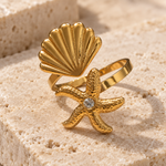 A gold-toned ring featuring a scalloped shell top and a starfish-shaped band with a clear rhinestone center, displayed on a textured beige stone surface.