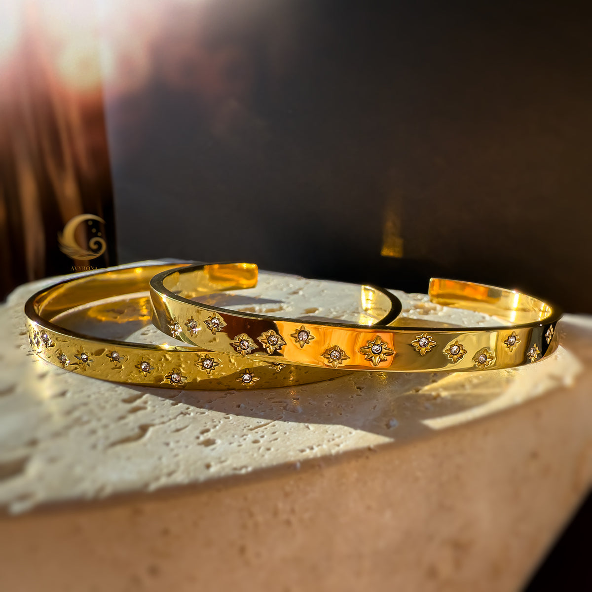 Golden starry cuff bracelets placed on textured beige stone, glowing in a dreamy sunset ambiance.