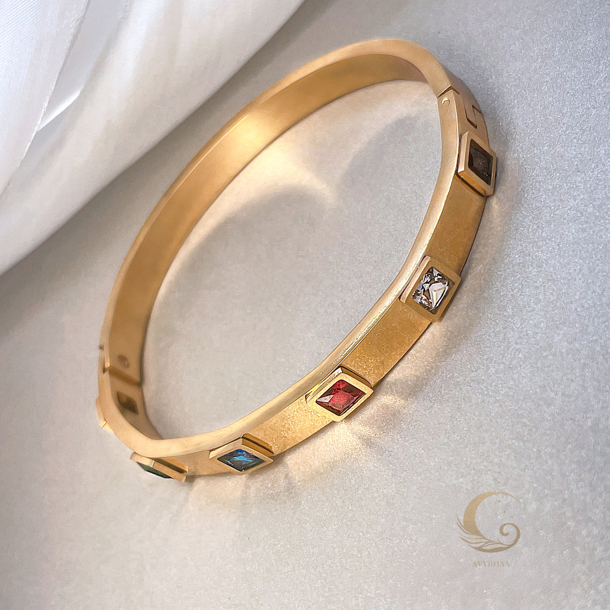 Bold gold bangle with vibrant red, clear, blue, and dark crystal accents, side view on neutral background – Fire Collection – Avyrona