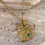 Gold pendant with a turquoise stone on a gold chain against a beige stone background