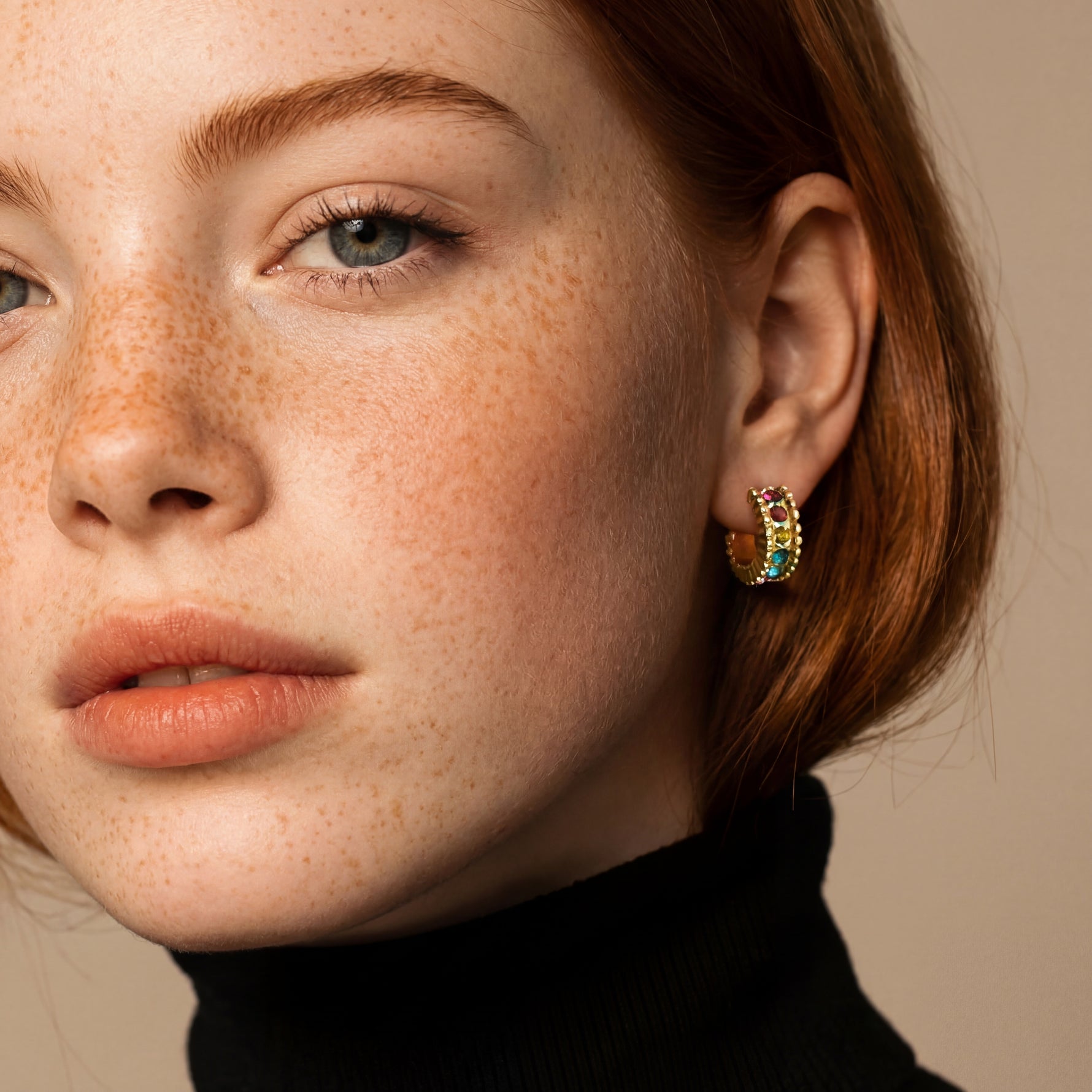 Close-up of a person wearing gold hoop earrings with gemstones on a beige background