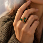 A close-up of a person's hand wearing two gold rings with oval green gemstones, set against a soft-focus background of brown knit clothing and warm sunlight.