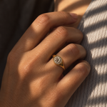 A gold signet ring with a circular sunburst design set with clear stones, worn on a person's hand resting against a textured knit garment.
