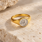 A gold signet ring with a silver-toned crescent moon and sun face detailing, accented with small clear stones, resting on a textured beige stone surface.