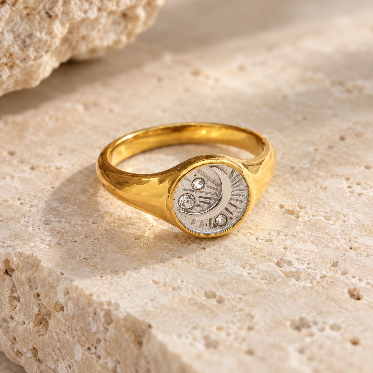 A gold signet ring with a silver-toned crescent moon and sun face detailing, accented with small clear stones, resting on a textured beige stone surface.