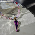 A beaded necklace with a glossy purple teardrop pendant, featuring pink, lavender, and white beads with small gold spacer beads, displayed on a light marble slab in bright natural light.