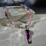 A beaded necklace with pink, lavender, white, and red beads, featuring a metallic purple teardrop-shaped pendant with gold accents, displayed on a marble block with dramatic sunlight and shadows.
