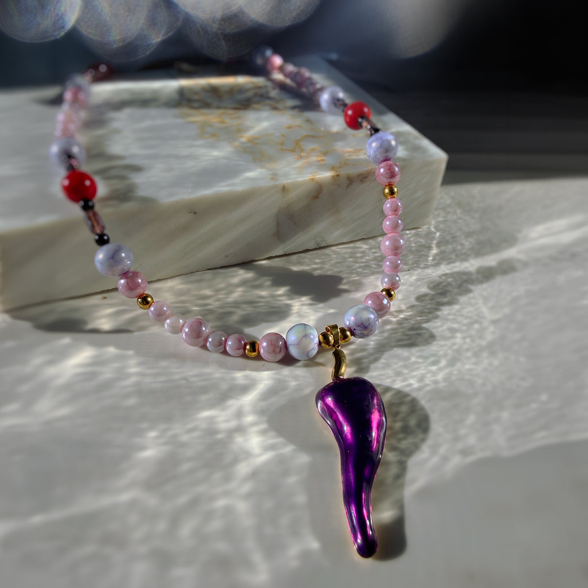 A beaded necklace with pink, lavender, white, and red beads, featuring a metallic purple teardrop-shaped pendant with gold accents, displayed on a marble block with dramatic sunlight and shadows.
