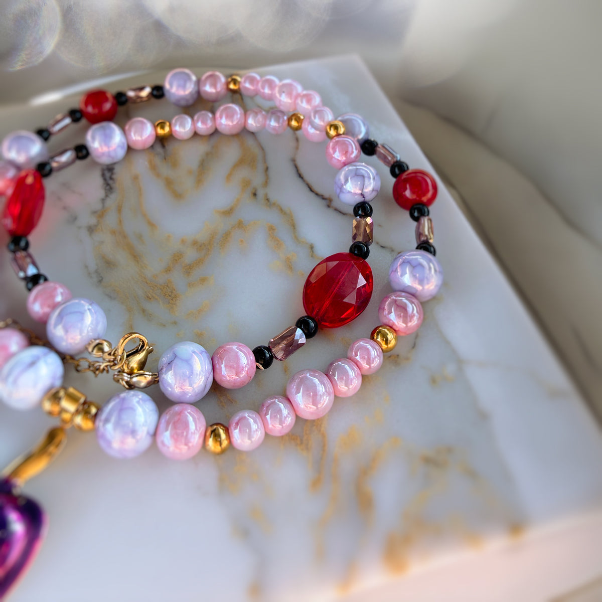 A close-up of a beaded strand jewelry piece resting on a white marble surface. The strand features iridescent light pink and lavender pearls, translucent red faceted beads, small gold spacer beads, and black accent beads.
