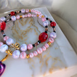A close-up of a beaded strand jewelry piece resting on a white marble surface. The strand features iridescent light pink and lavender pearls, translucent red faceted beads, small gold spacer beads, and black accent beads.