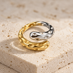 A two-tone metallic ring with a wavy, organic design, featuring one half in polished gold and the other in polished silver, resting on a textured beige stone surface.