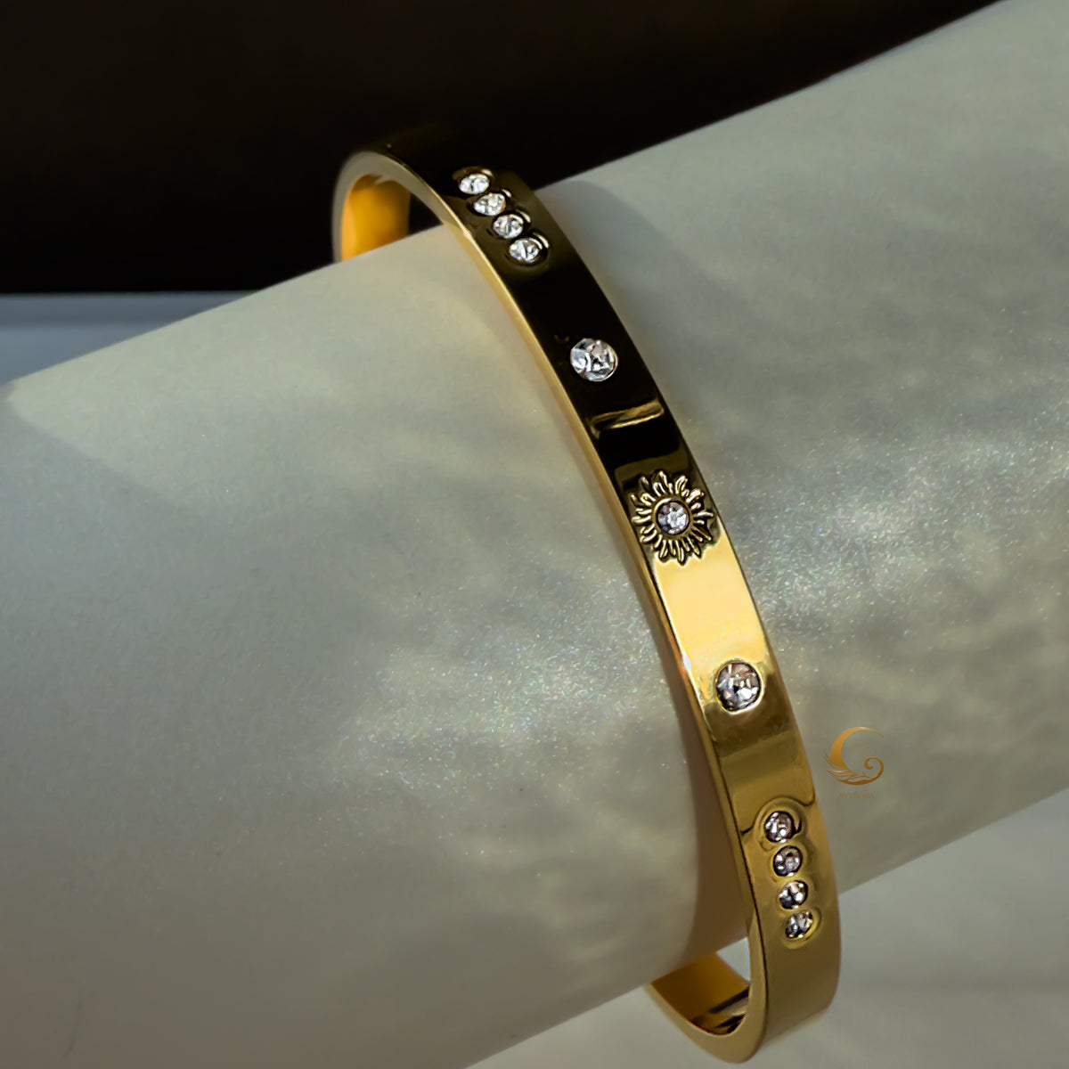 A gold bracelet with a sun motif and small cubic zirconia stones, displayed on a smooth, light-colored tube.