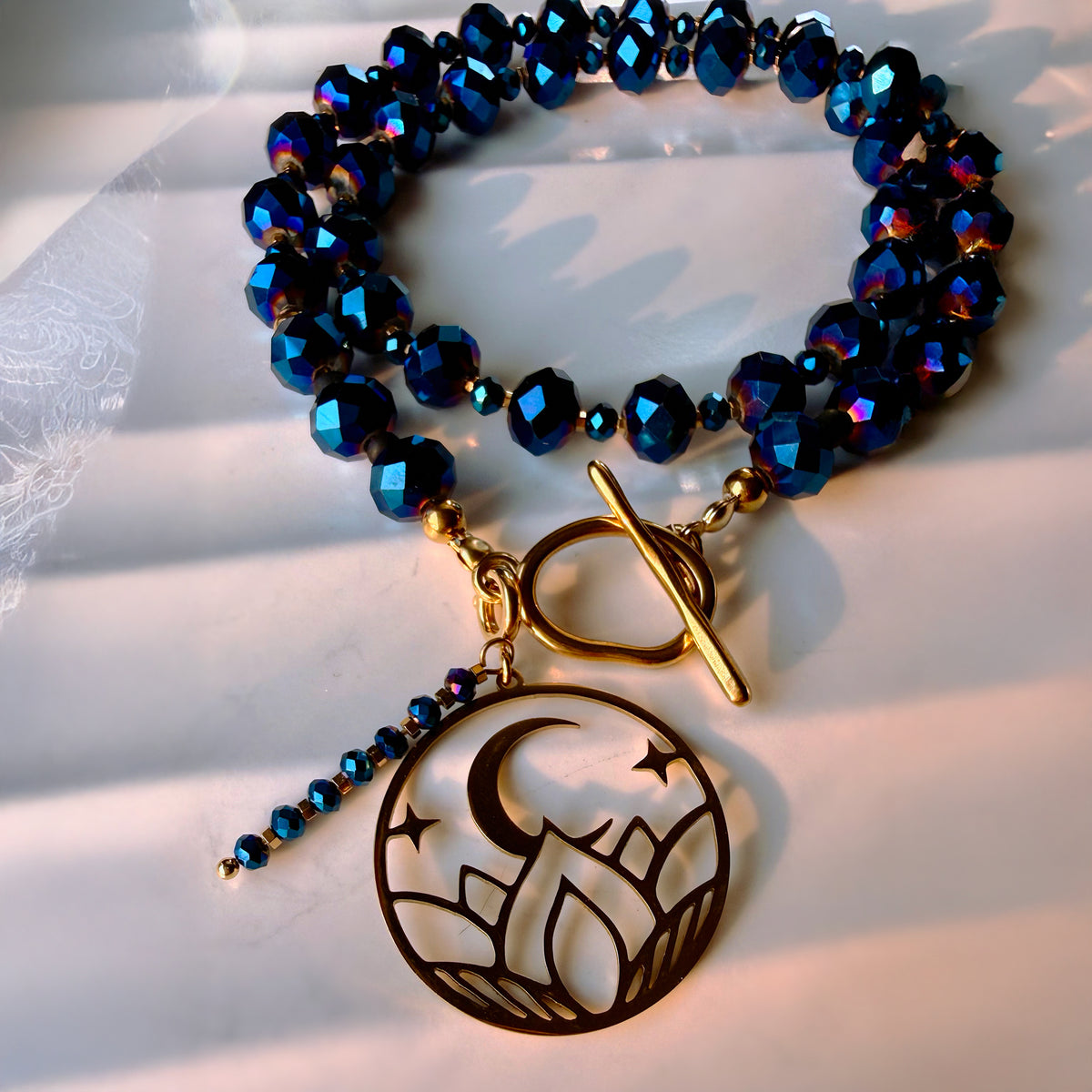A beaded bracelet with dark blue faceted beads, gold-toned toggle clasp, and a dangling circular charm featuring a crescent moon and stars over mountain-like shapes, photographed on a white surface with soft shadows.