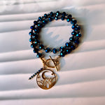 A beaded stretch bracelet with dark blue faceted beads and a gold-toned toggle clasp, featuring a dangling crescent moon and stars charm, displayed on a white slatted surface.