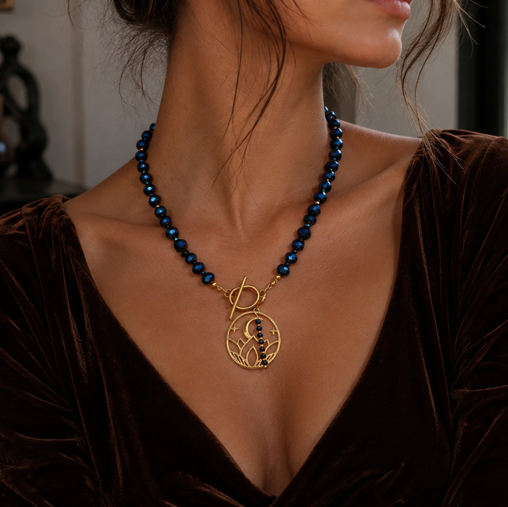 A close-up of a woman wearing a dark blue beaded necklace with a gold pendant featuring a stylized letter 'S' and small dark blue stones, paired with a brown velvet top.