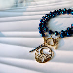 A beaded bracelet with deep blue faceted beads and a gold-toned toggle clasp, featuring a circular moon and star pendant and a small matching bead strand, photographed on a white draped fabric background.