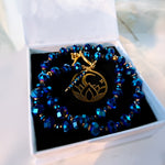 A beaded bracelet with faceted blue and iridescent beads, a gold toggle clasp, and a central gold lotus pendant, displayed inside an open white jewelry box with a black insert.
