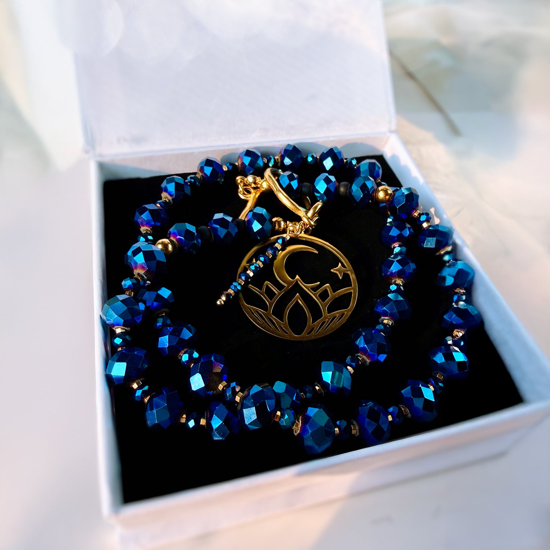 A beaded bracelet with faceted blue and iridescent beads, a gold toggle clasp, and a central gold lotus pendant, displayed inside an open white jewelry box with a black insert.