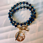 A beaded bracelet with faceted dark blue beads, gold-toned spacer beads, a toggle clasp, and a dangling circular charm featuring a flame motif and small blue beads.