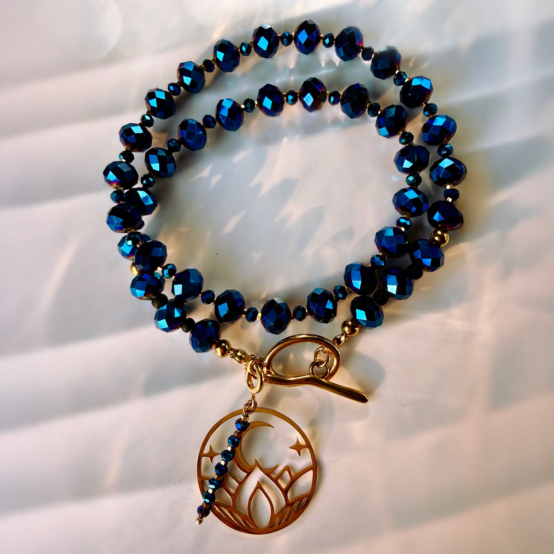 A beaded bracelet with faceted dark blue beads, gold-toned spacer beads, a toggle clasp, and a dangling circular charm featuring a flame motif and small blue beads.