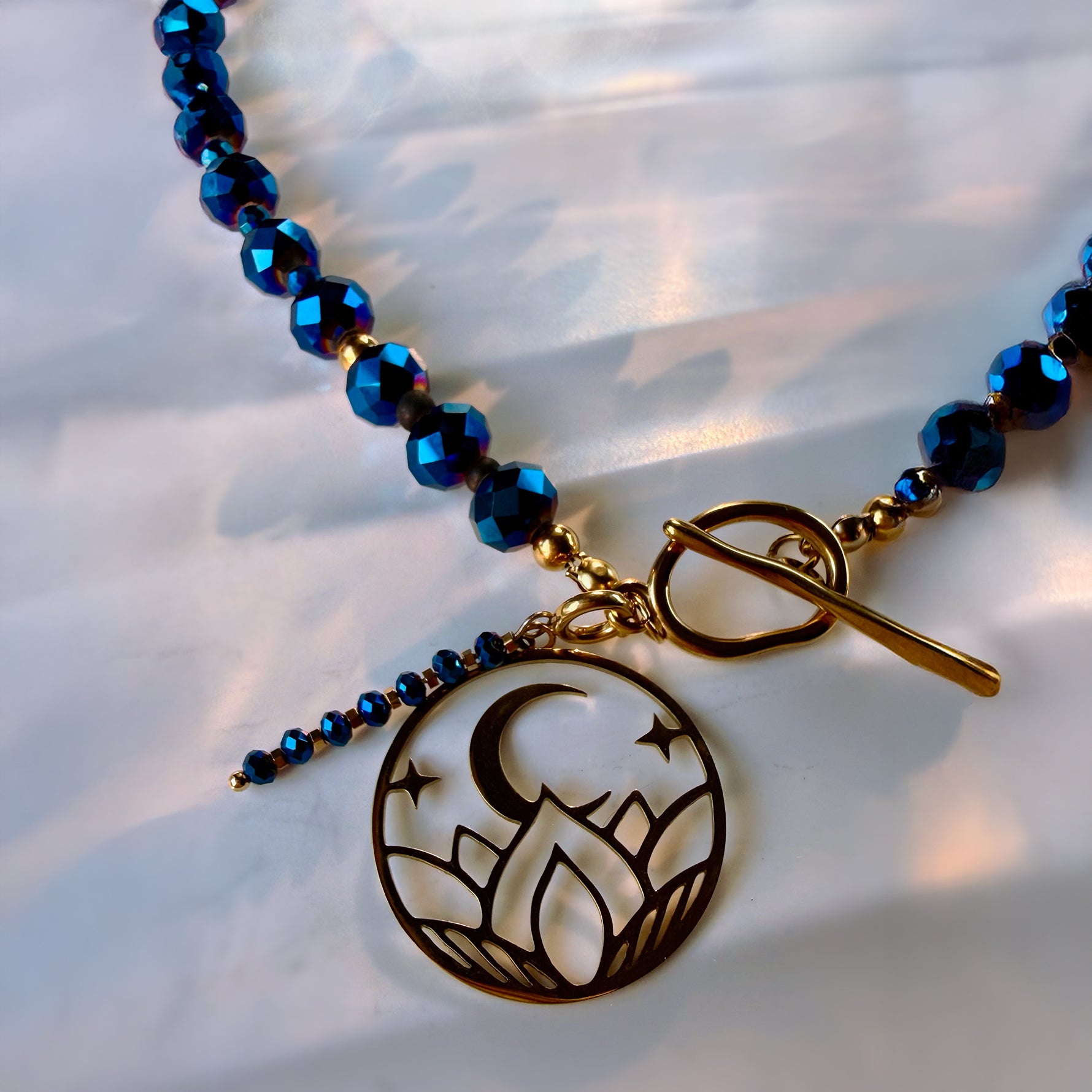 A close-up of a beaded necklace with faceted dark blue beads, a gold toggle clasp, and a circular pendant featuring a crescent moon and mountain design over a lotus flower, displayed on a white textured fabric background.