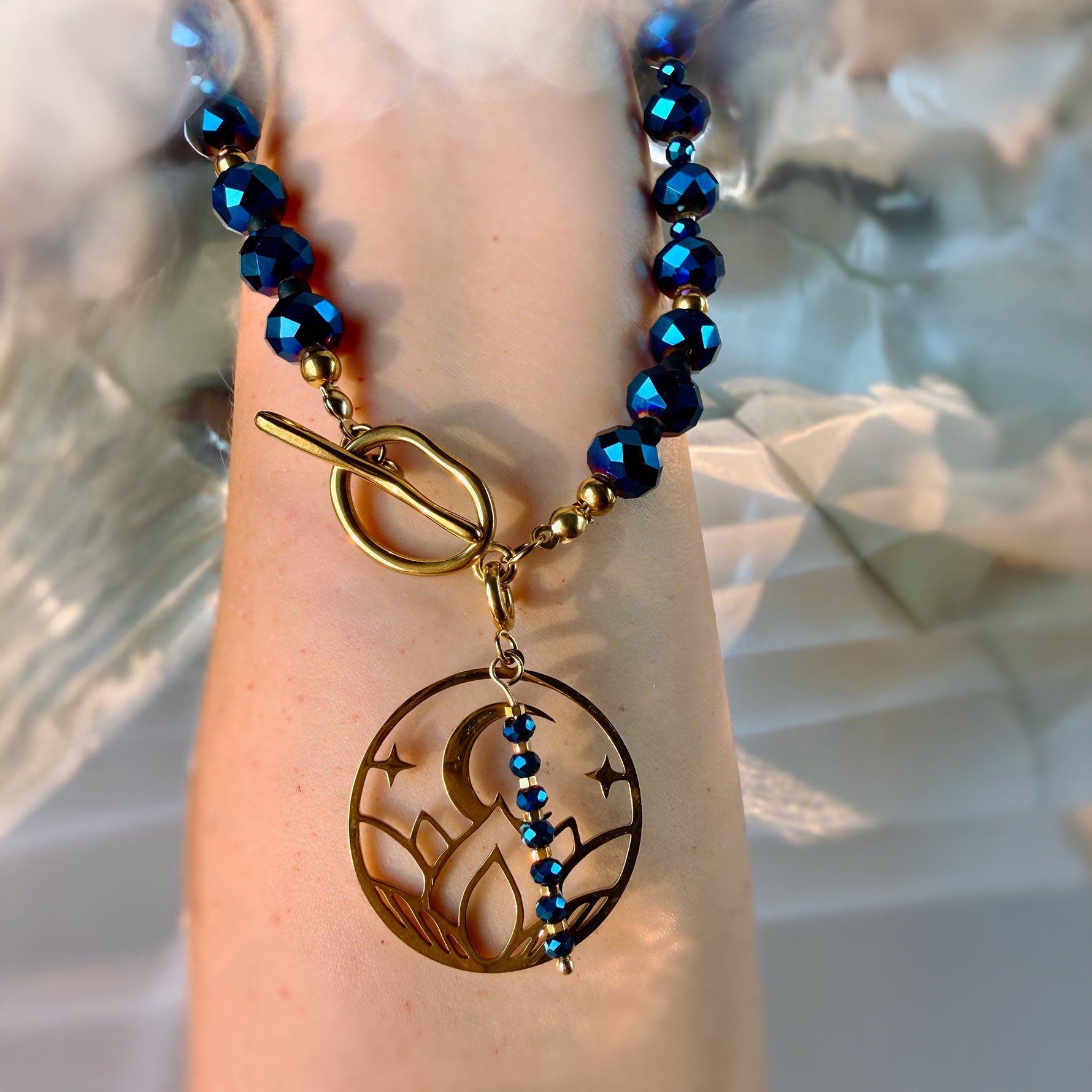 A close-up of a beaded bracelet with metallic blue faceted beads and a gold toggle clasp, worn on a person's wrist. The bracelet features a circular gold pendant with a crescent moon and star design, accented by a vertical line of small blue beads.