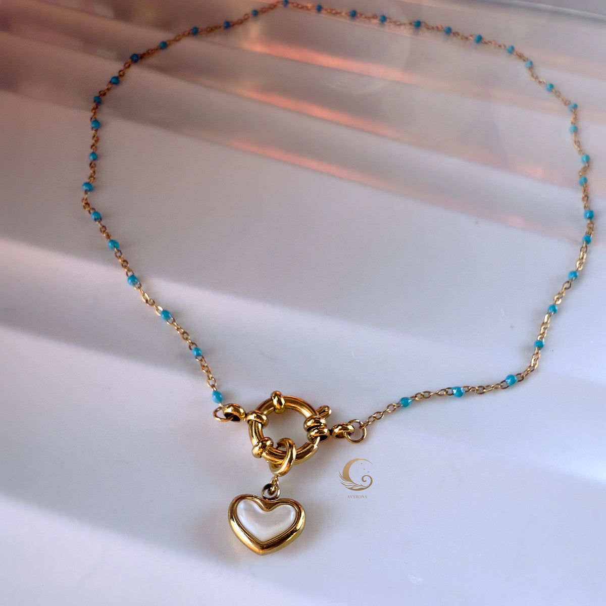 The Alia necklace arranged on a flat surface in a soft arc, showing the shimmering gold heart charm and toggle clasp, with light blue beads glowing under warm sunset-inspired lighting.