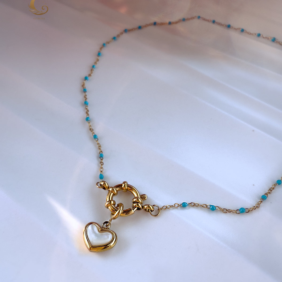 Close-up of the Alia necklace laid flat in a circle on a white background, showing delicate light blue beads, a gold toggle clasp, and a dangling heart-shaped mother of pearl pendant.