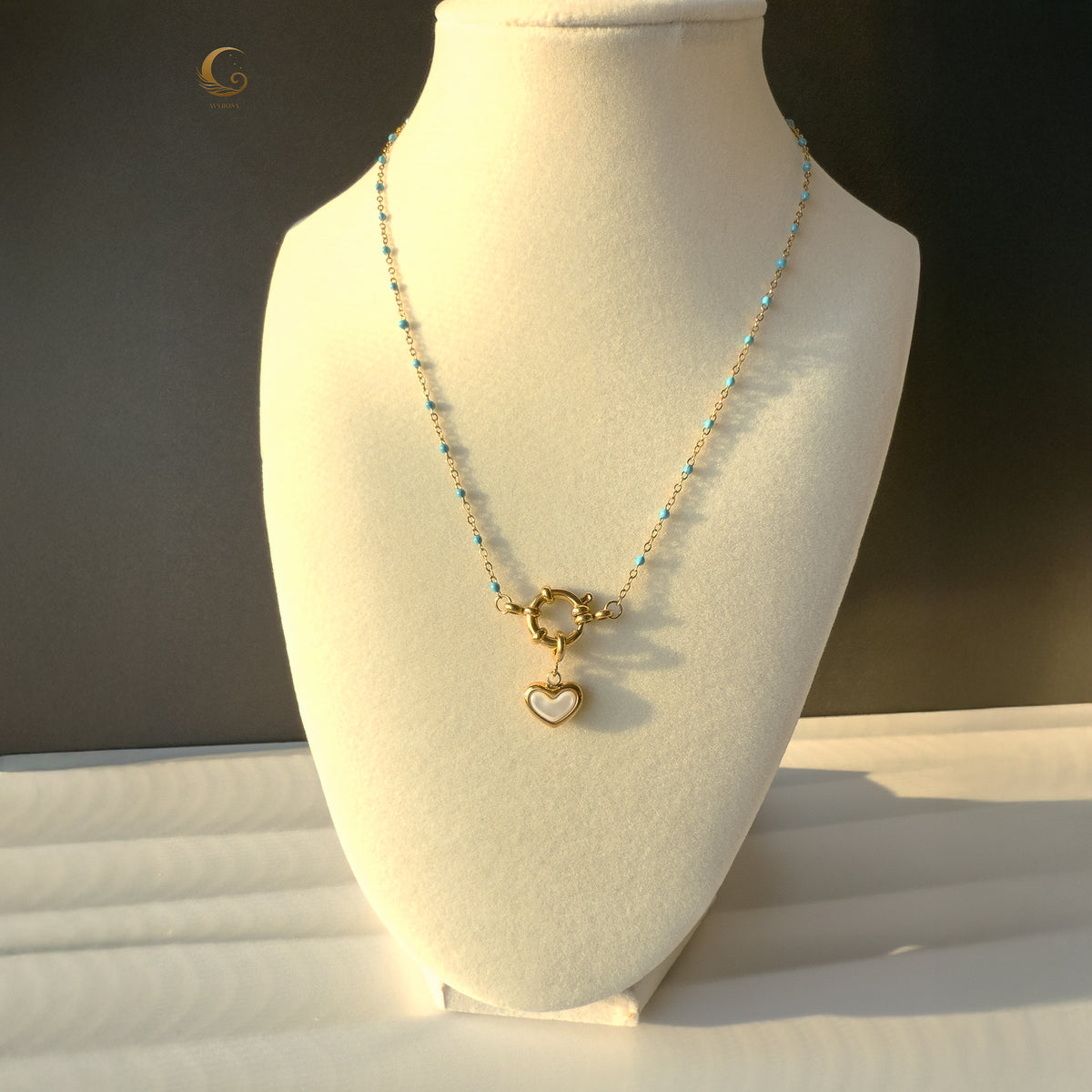 The Alia necklace displayed on a white jewelry bust, highlighting its soft blue beaded chain and central gold heart charm made of mother of pearl, photographed in natural daylight.