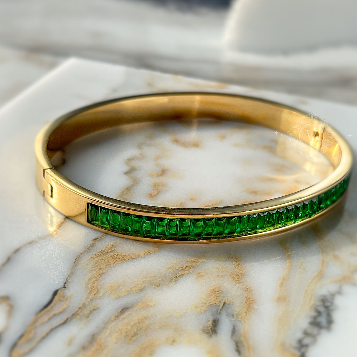 Full view of gold bangle bracelet with vibrant green crystals, placed on marble background – elegant and powerful look