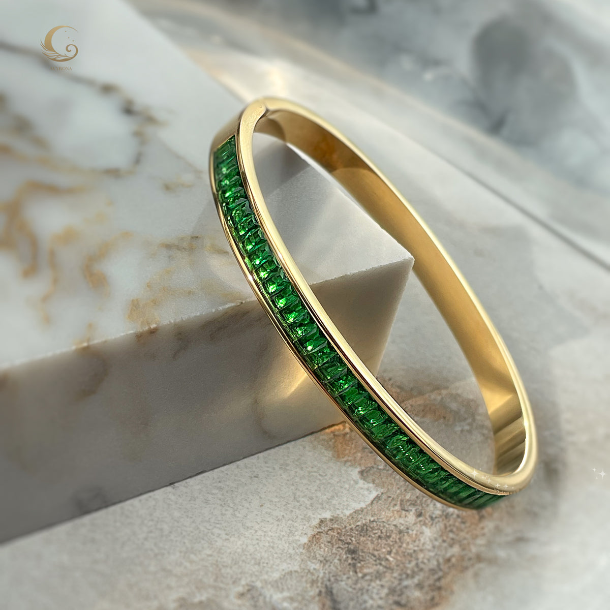 Gold cuff bracelet with emerald green crystals leaning on marble block – luminous Fire Collection jewelry by Avyrona