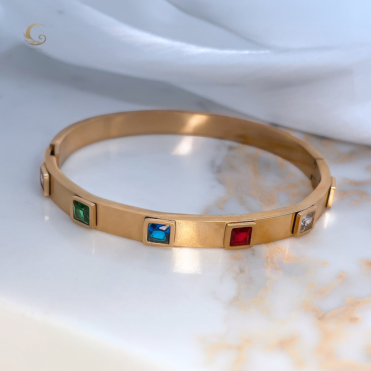 Colorful crystal gold bracelet with red, blue, green, and clear stones displayed on satin fabric – Fire-inspired jewelry by Avyrona