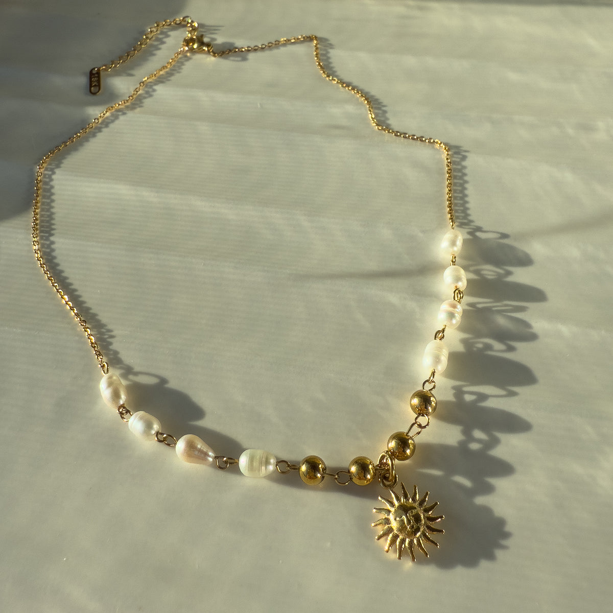 Ayla necklace laid out flat on white surface, showing gold beads, pearl accents, and sun charm.