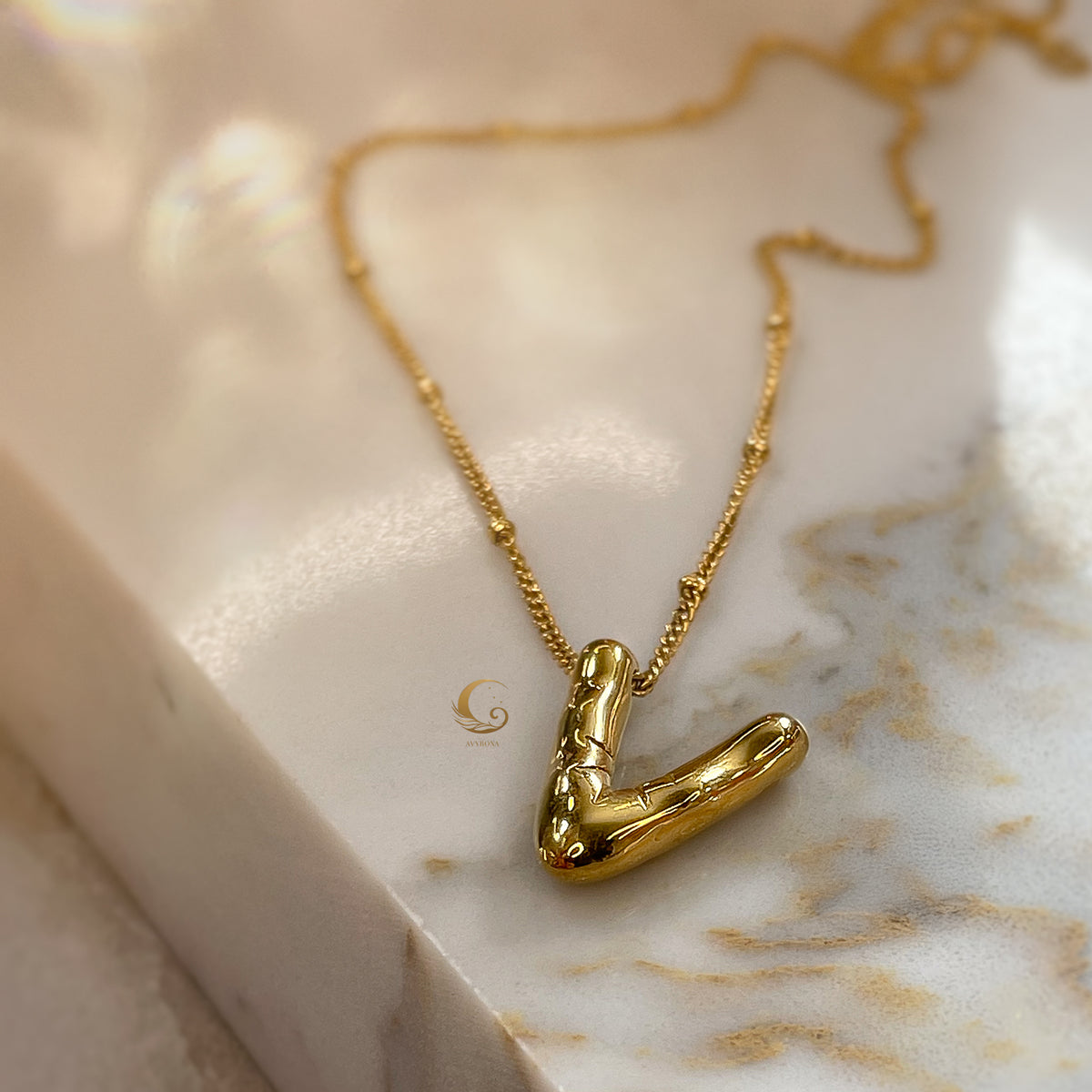 Single gold balloon-style letter “V” necklace on satellite chain, resting on soft beige marble.