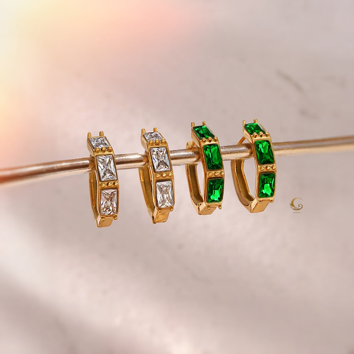 Four Camila hoop earrings displayed on a gold rod, showing two emerald green and two clear zircon stone variants