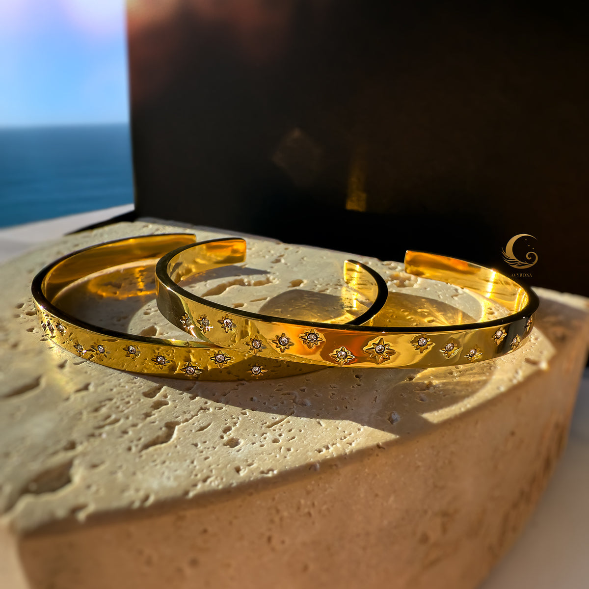 Two gold cuff bracelets with celestial star engravings and cubic zirconia, resting on a natural stone under warm light.
